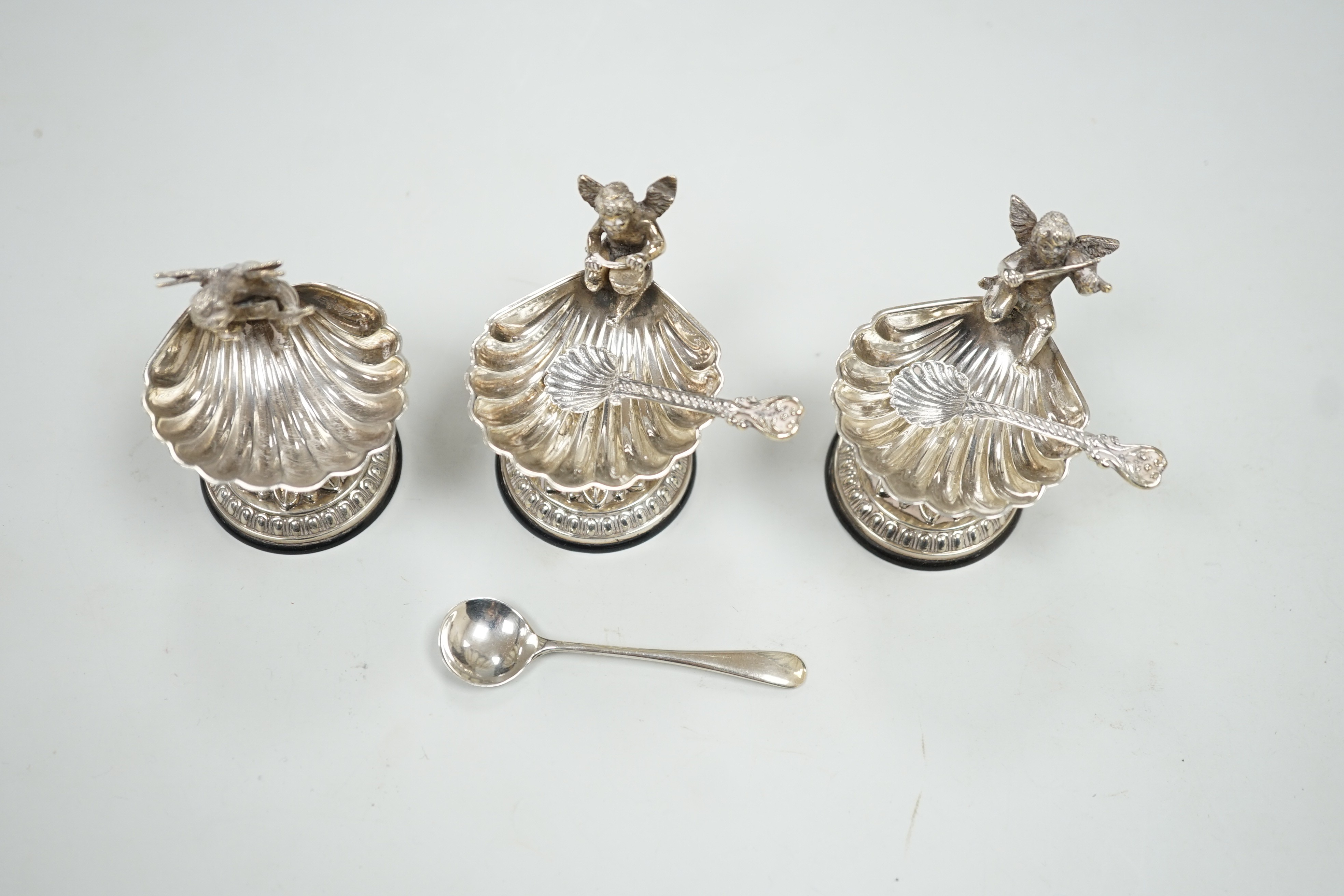 A set of three 20th century Spanish white metal shell pedestal salts with cherub handles and three spoons, height 50mm.